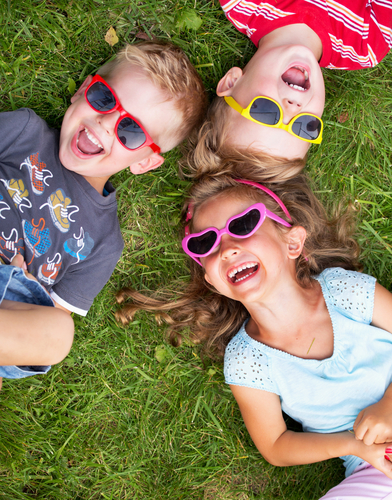 Billede af 3 små børn med solbriller på som ligger i græsset og griner