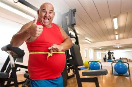 Glad, overvægtig mand i rød trøje holder gult målebånd om maven i træningscenter.
